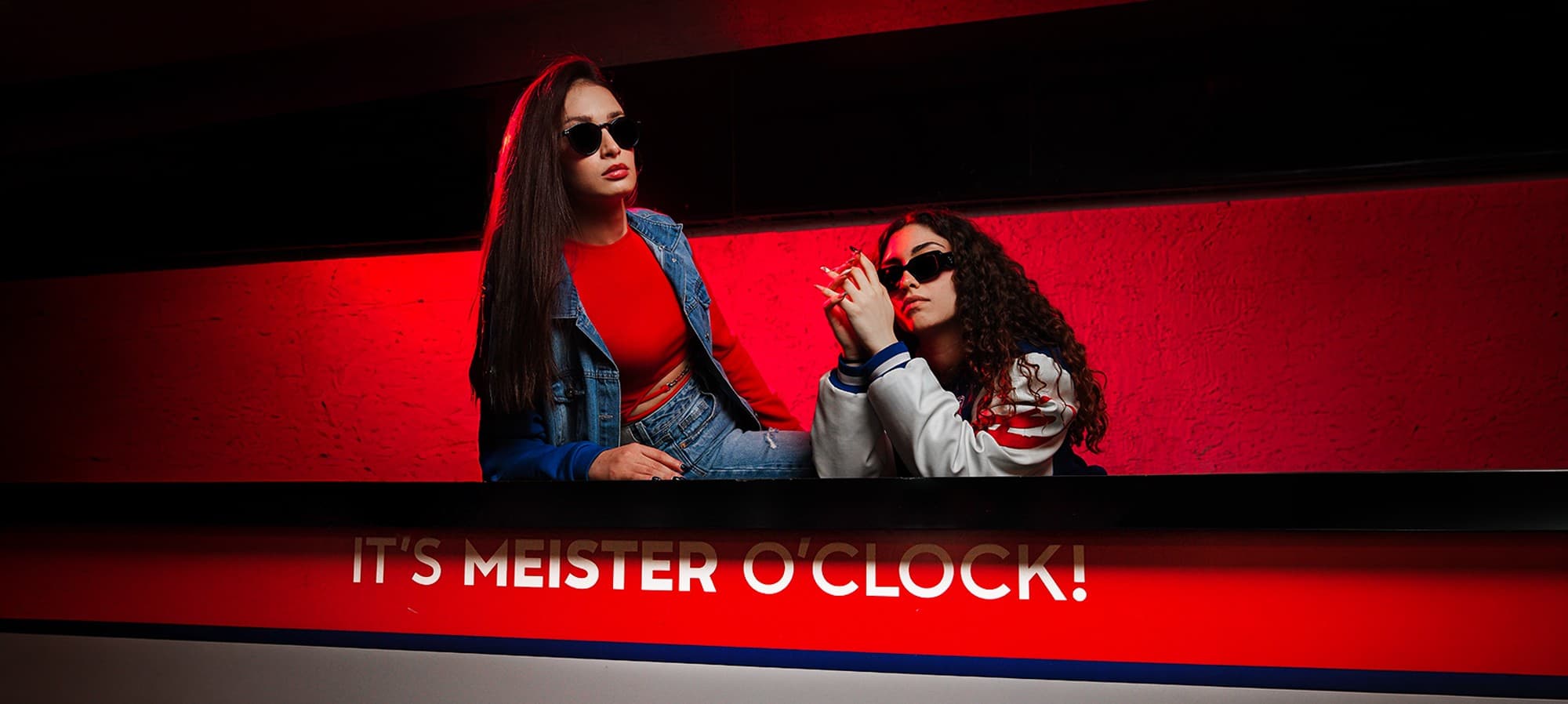 Kırmızı ışıklı tezgâhta güneş gözlüklü iki arkadaş; tezgâhta 'It's Meister O'Clock!' yazısı